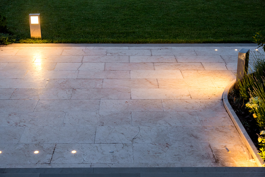 The pavement of marble tiles and a stone border area in the evening illuminated by ground lanterns in a metal housing shining a warm light around the lawn.